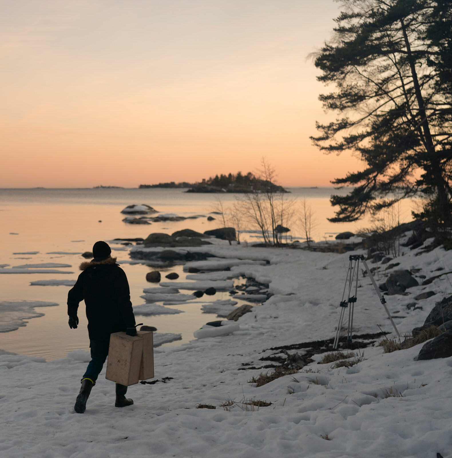 Henkilö kävelee lumisella rannalla auringonlaskun aikaan kantaen laatikkoa. Taustalla näkyy jäinen meri ja kivinen rantaviiva