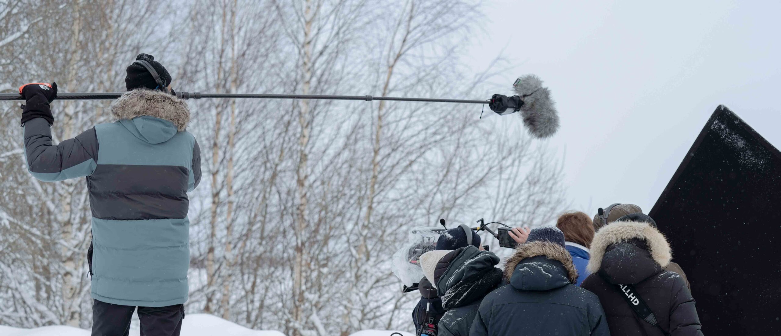 Kuvausryhmä työskentelee lumisessa ympäristössä, jossa yksi henkilö pitelee puomimikrofonia ja muut huolehtivat kamerasta ja valolaitteista.